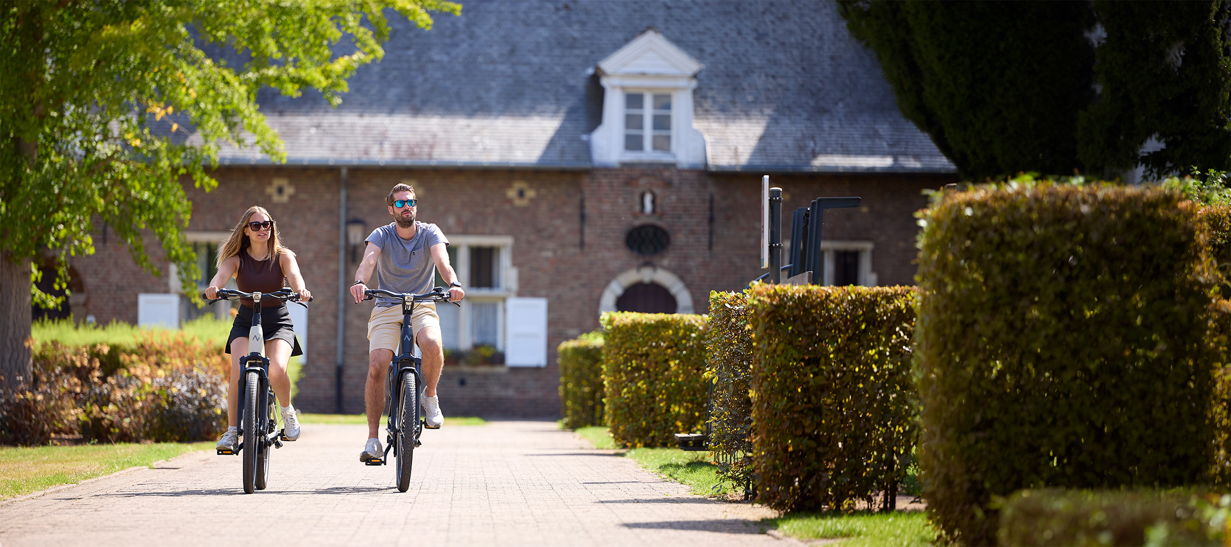 Twee mensen fietsen op elektrische fietsen in een groene, historische omgeving.