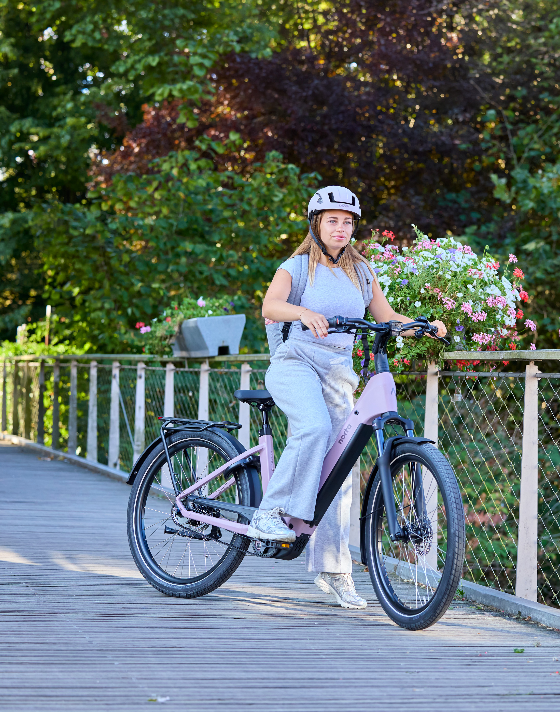 Vrouw staat naast haar elektrische fiets van Norta op een brug met bloemen. De fiets heeft de kleur pastel violet.