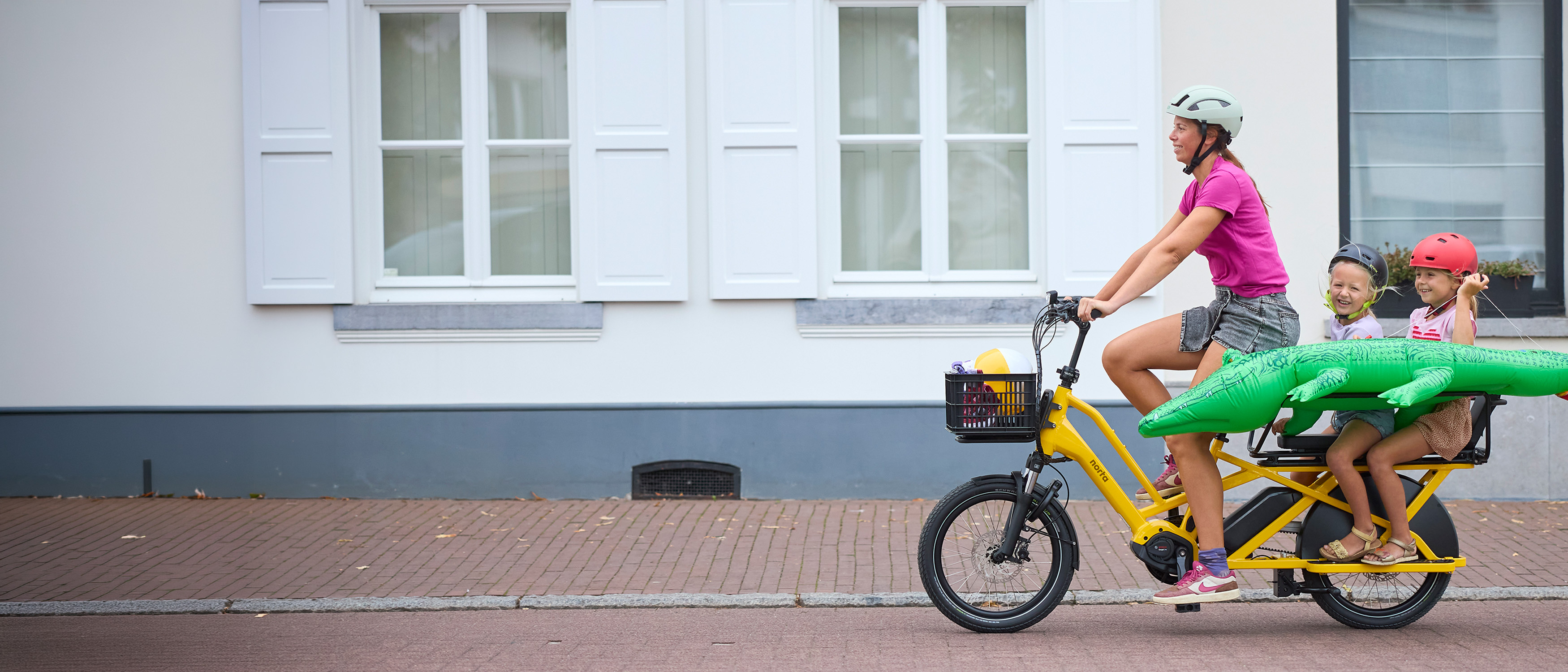 Vrouw met twee kinderen fietsen met een gele elektrische longtail door het dorp op weg naar de zee.