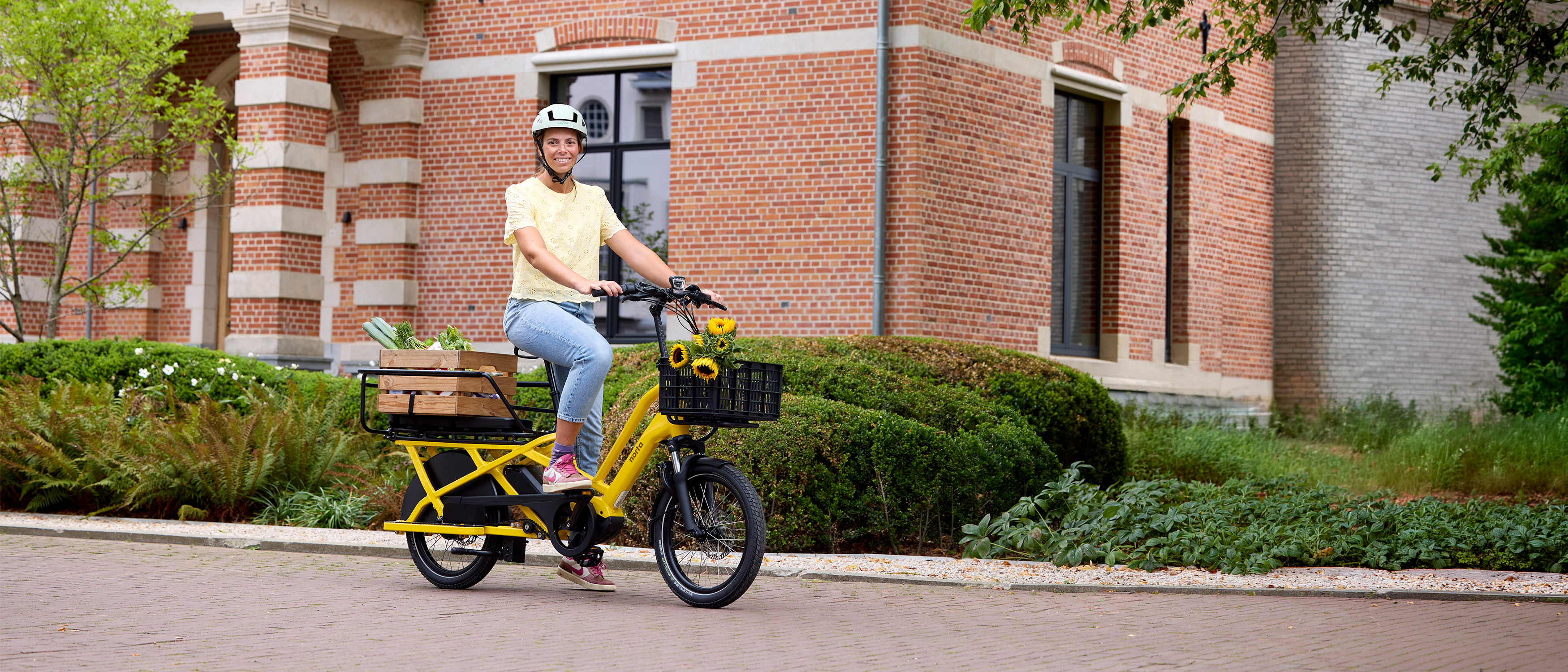 Vrouw zit op een gele cargobike van Norta. De mand is gevuld met zonnebloemen.